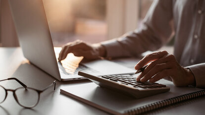 Mensch arbeitet am Tisch mit Laptop und Taschenrechner